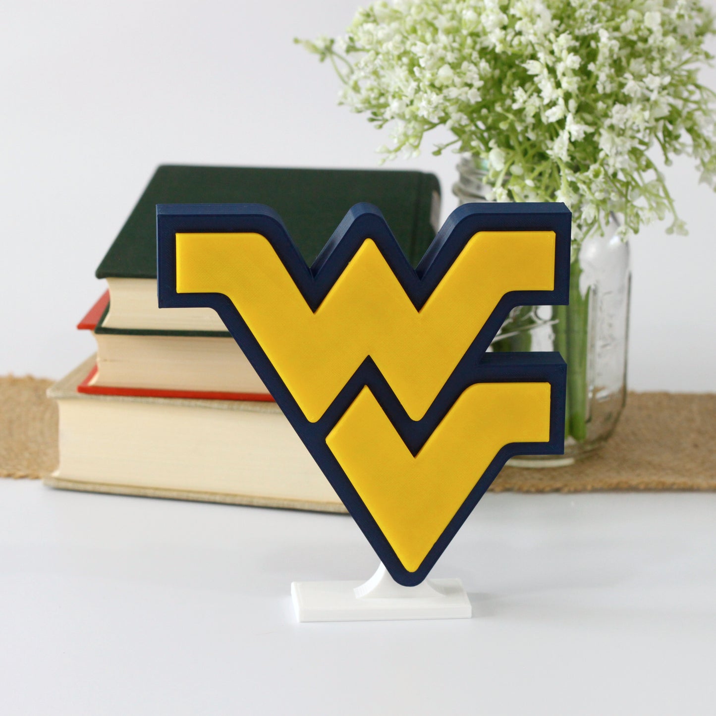 West Virginia University decoration in front of a stack of books and mason jar with baby's breath flowers.